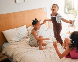 Three young children hold hands while they jump on a bed.