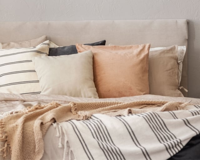 A cozy bed with neutral-colored throw pillows and blankets next to a vase of flowers on a bedside table.
