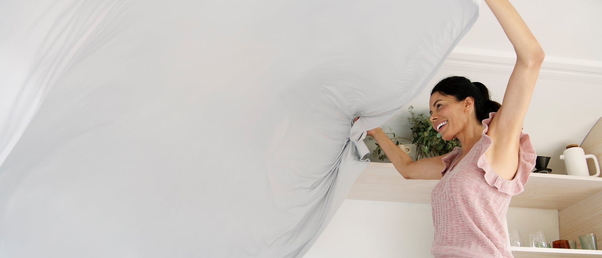 A smiling woman waves a sheet in the air while putting it on a bed.